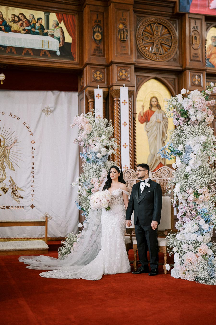 Bride and groom standing in an ornate church setting, surrounded by floral arrangements, capturing a wedding moment with religious artwork in the background.