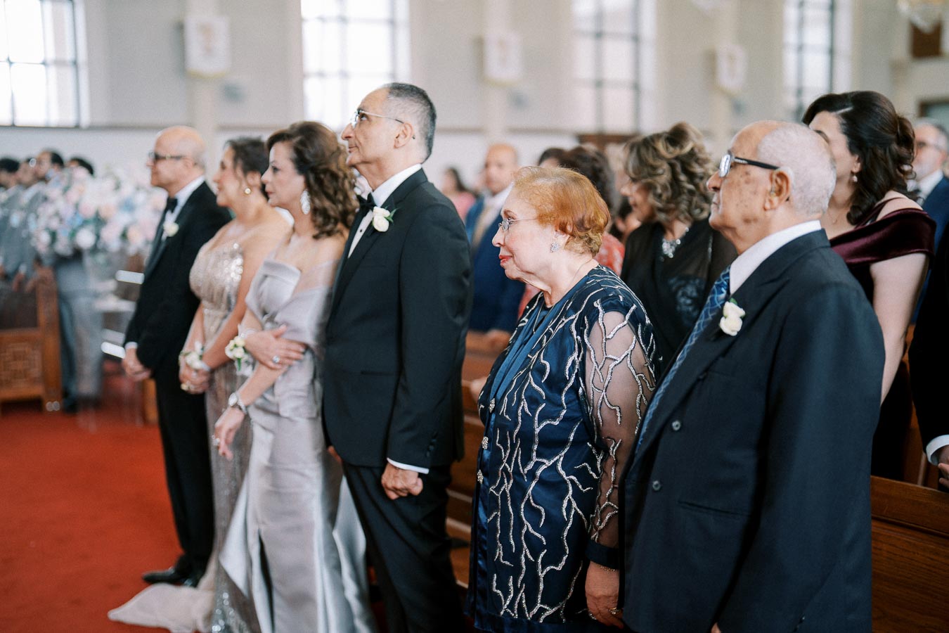 A group of elegantly dressed people standing in a church during a wedding ceremony, with some wearing formal suits and others in evening gowns, attentively observing the event.