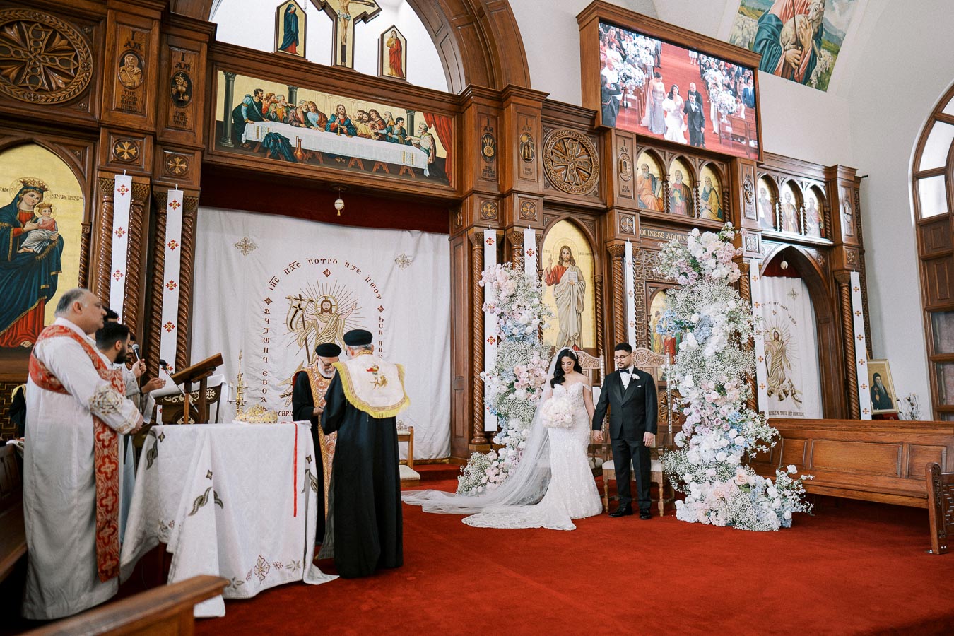 Orthodox wedding ceremony inside a beautifully decorated church, featuring a bride and groom standing near the altar adorned with floral arrangements and ornate religious icons.