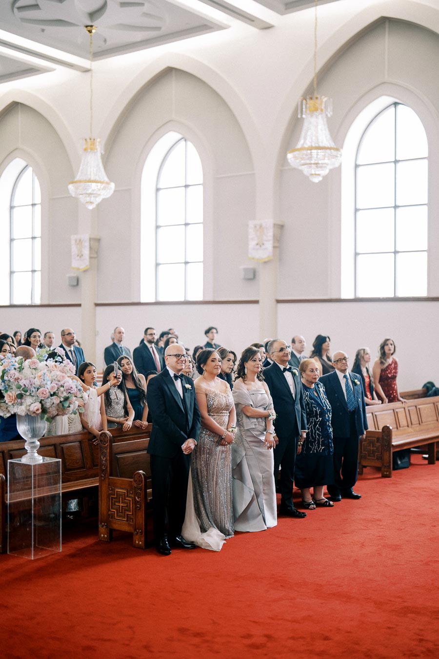 A gathering of elegantly dressed guests inside a beautifully decorated church with high ceilings and chandeliers, attending a formal ceremony.