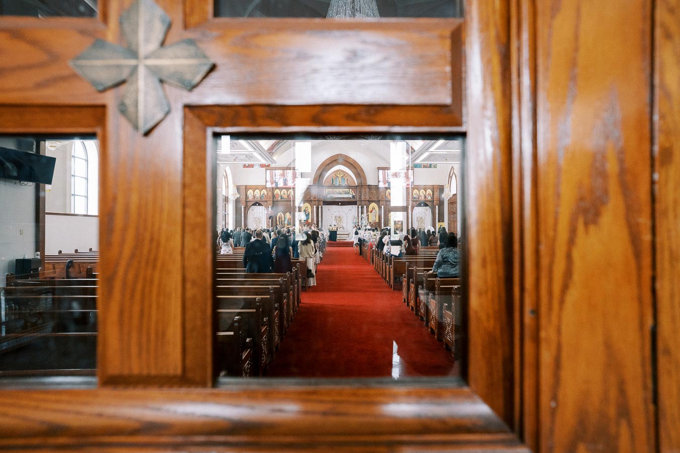 View through a wooden church door window, showing an interior with people attending a service, red carpet aisle, and ornate altar in the background, highlighting the church's architecture and communal gathering.