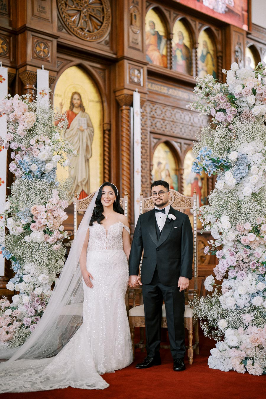 A bride in a lace wedding gown and a groom in a black tuxedo stand hand in hand in a beautifully decorated church with ornate wooden carvings and floral arrangements, capturing an elegant and traditional wedding ceremony.
