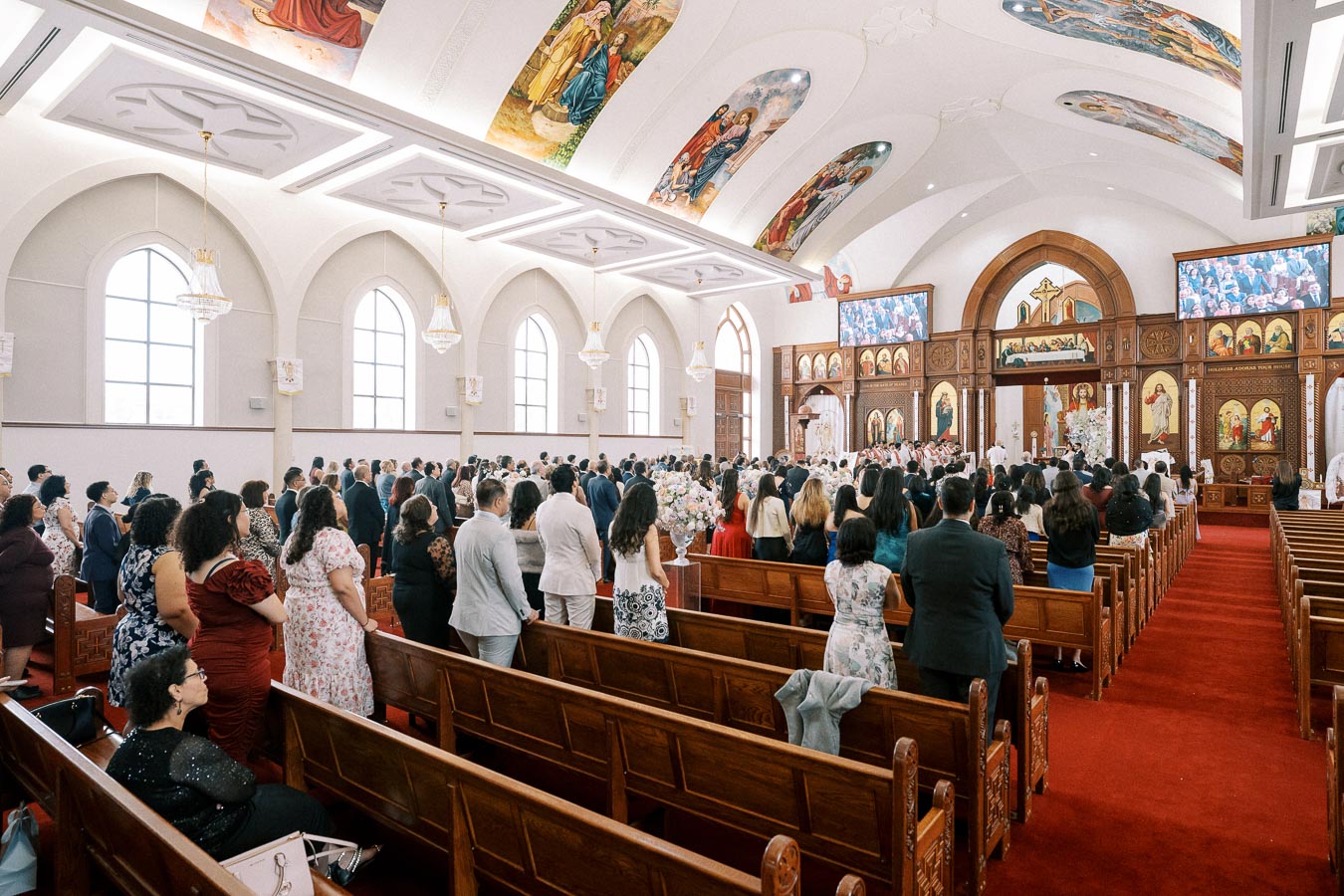 Alt text: Congregation attending a service in a large, ornate church with decorative murals on the ceiling and wooden pews. The interior is bright and spacious, featuring stained glass windows and chandeliers.