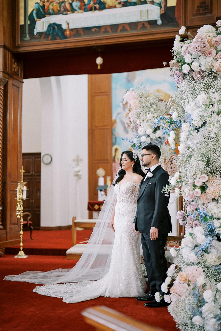 A bride and groom stand together at the altar of an ornately decorated church, surrounded by elegant floral arrangements. The bride wears a lace wedding dress with a long veil, and the groom is in a classic black tuxedo. The interior features red carpet and rich wood paneling, with a religious artwork overhead.