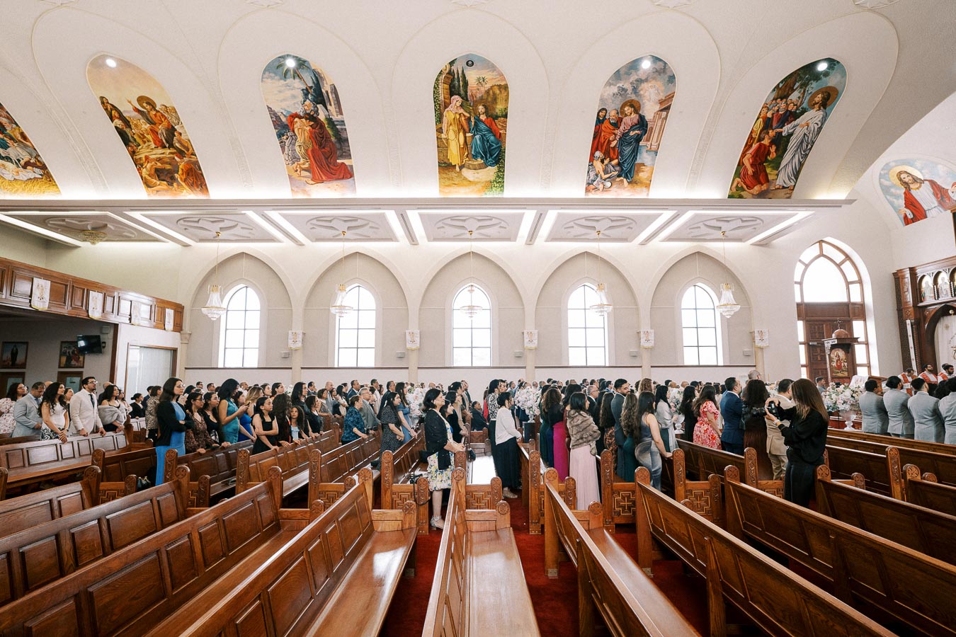Interior view of a church filled with people attending a formal event, featuring ornate wooden pews and arched windows. Beautiful religious murals adorn the ceiling, creating a spiritual and sacred atmosphere.