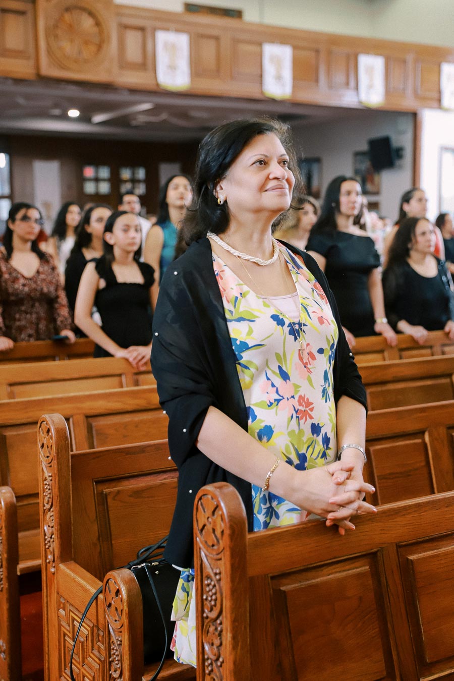 A woman in a floral dress and black shawl smiling while standing in a church, with other people in the background during a service.