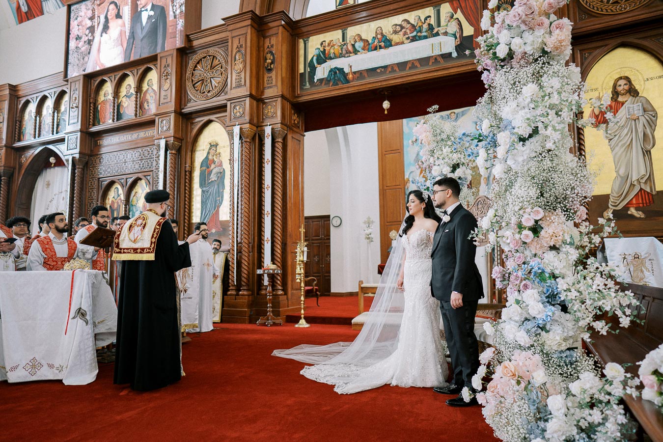 A bride and groom stand at the altar during a traditional church wedding ceremony, surrounded by ornate wooden architecture and floral decorations, with a priest conducting the service.