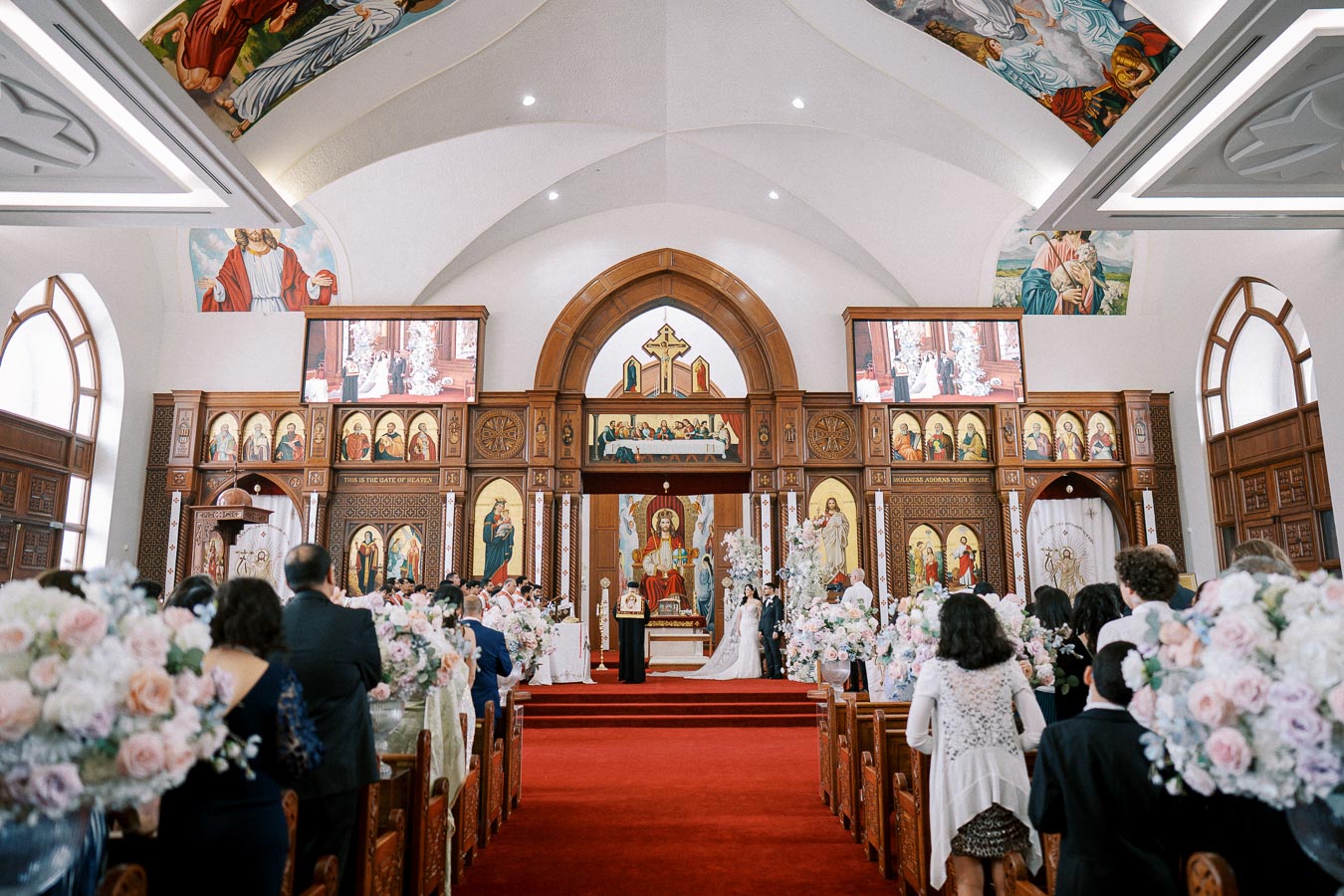Wedding ceremony in an ornate church with colorful stained glass windows and intricate religious artwork on the walls, showcasing a bride and groom at the altar surrounded by guests seated in pews adorned with floral arrangements.