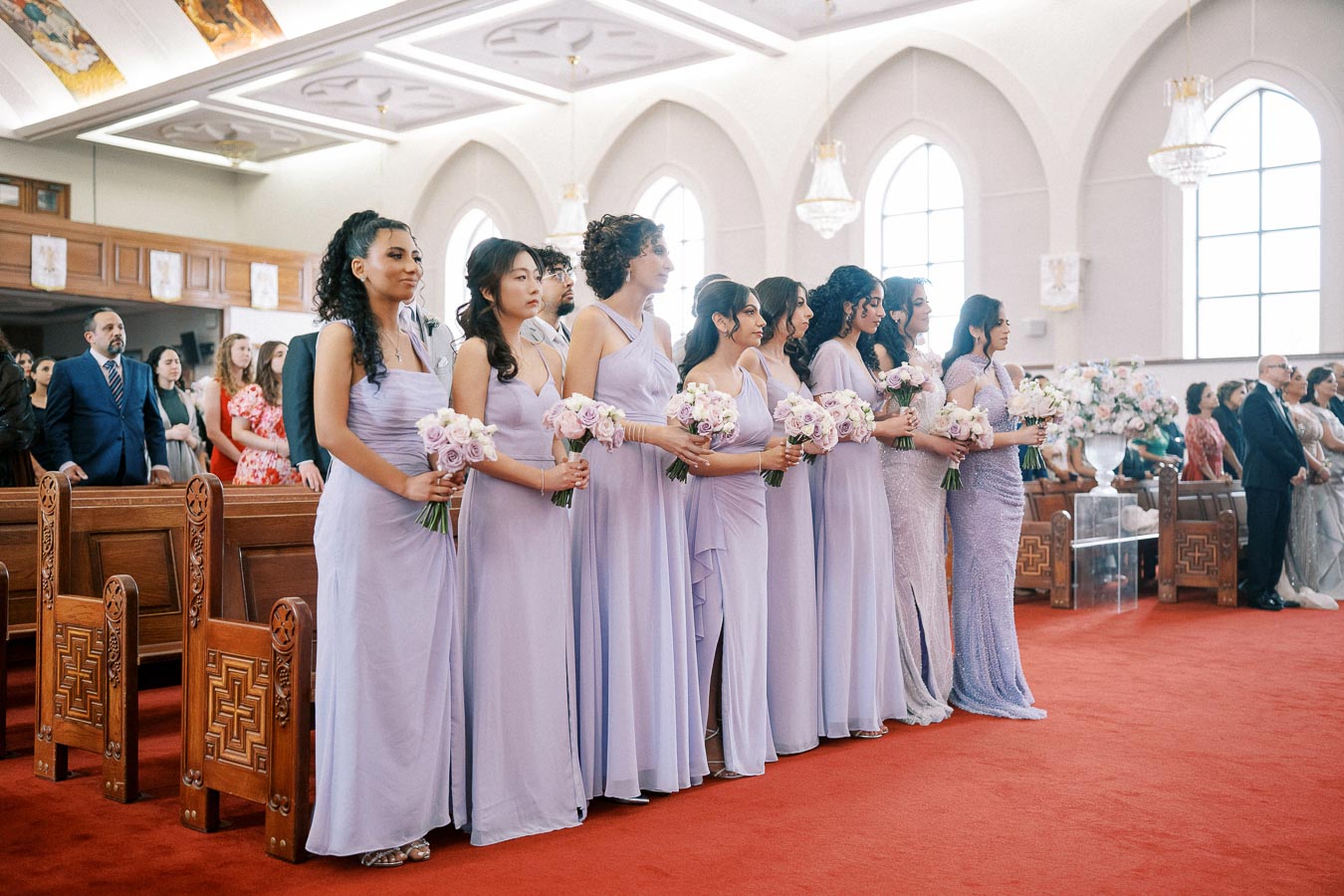 Bridesmaids in lavender dresses holding bouquets stand in a church during a wedding ceremony.