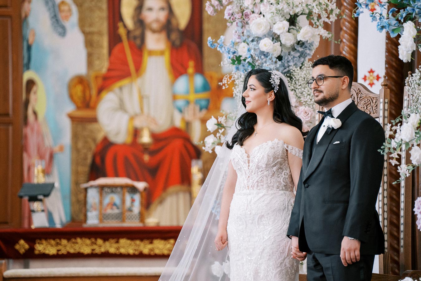 Alt text: Bride in a lace wedding dress and groom in a black tuxedo stand in a beautifully decorated church, with floral arrangements and religious artwork in the background.