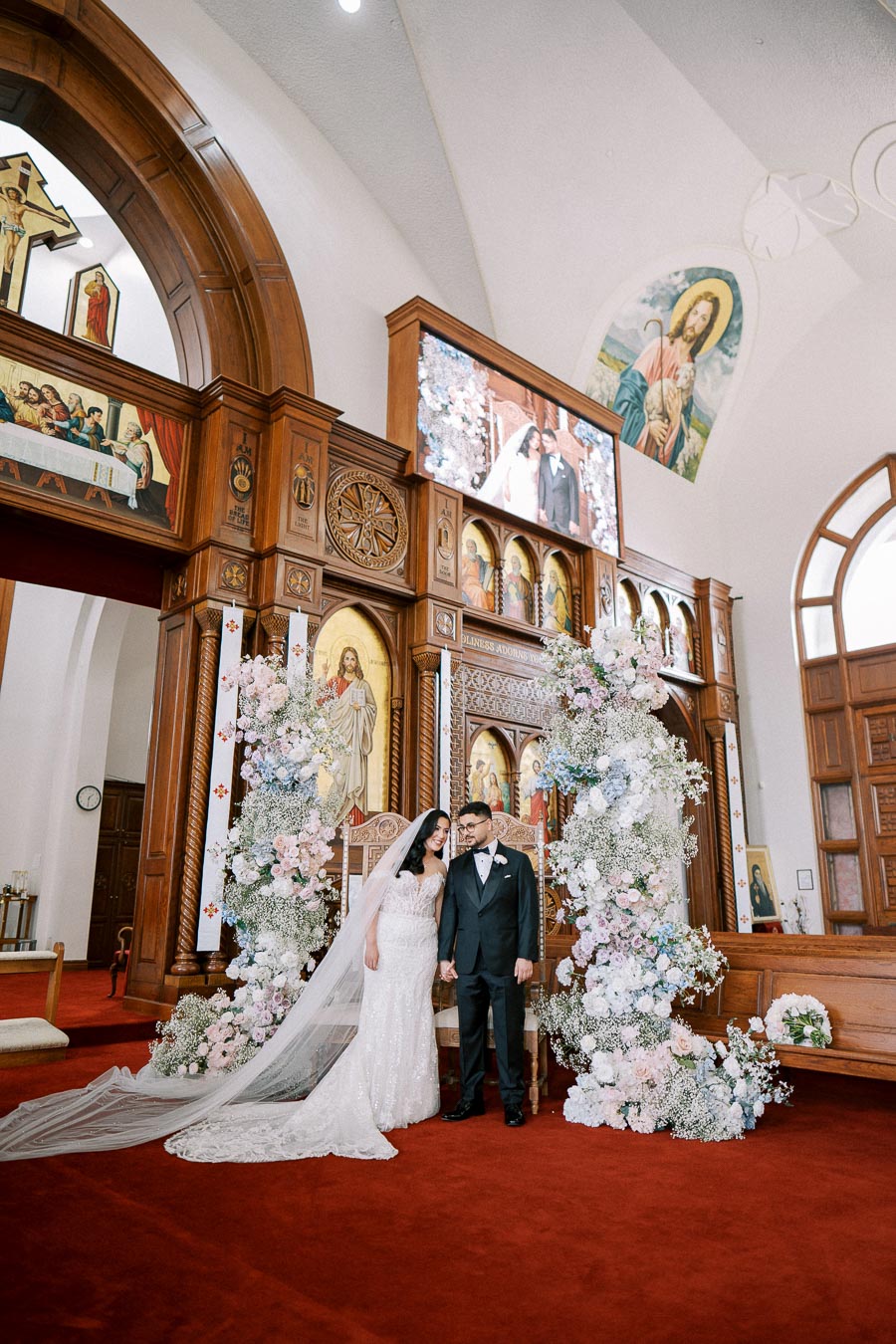 Elegant wedding ceremony in a beautifully decorated church interior with a bride in a lace gown and groom in a tuxedo standing under a floral arch, surrounded by religious artwork and stained glass windows.