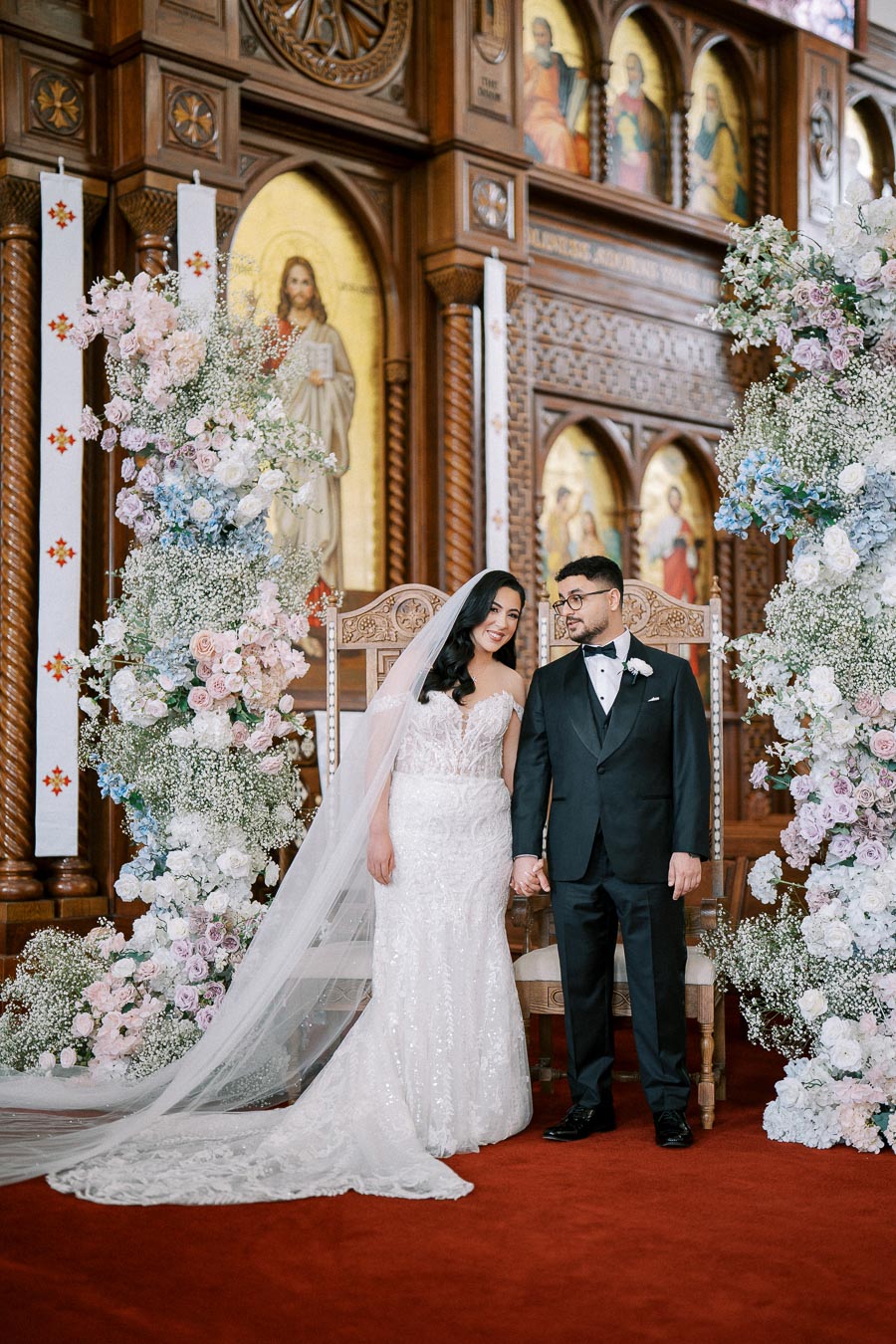 Wedding couple holding hands in a beautifully decorated church with ornate wooden carvings and floral arrangements, bride in a lace gown and groom in a black tuxedo.