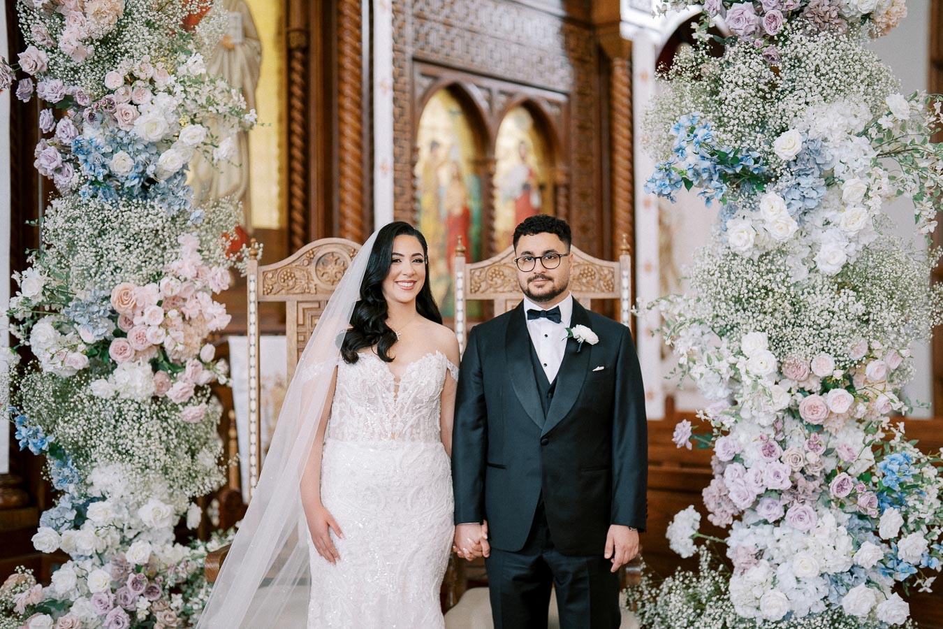 A bride and groom hold hands in a beautifully decorated church, with elegant floral arrangements in soft pastel colors surrounding them. The bride wears a stunning white lace gown and veil, while the groom looks sharp in a black tuxedo with a bow tie.