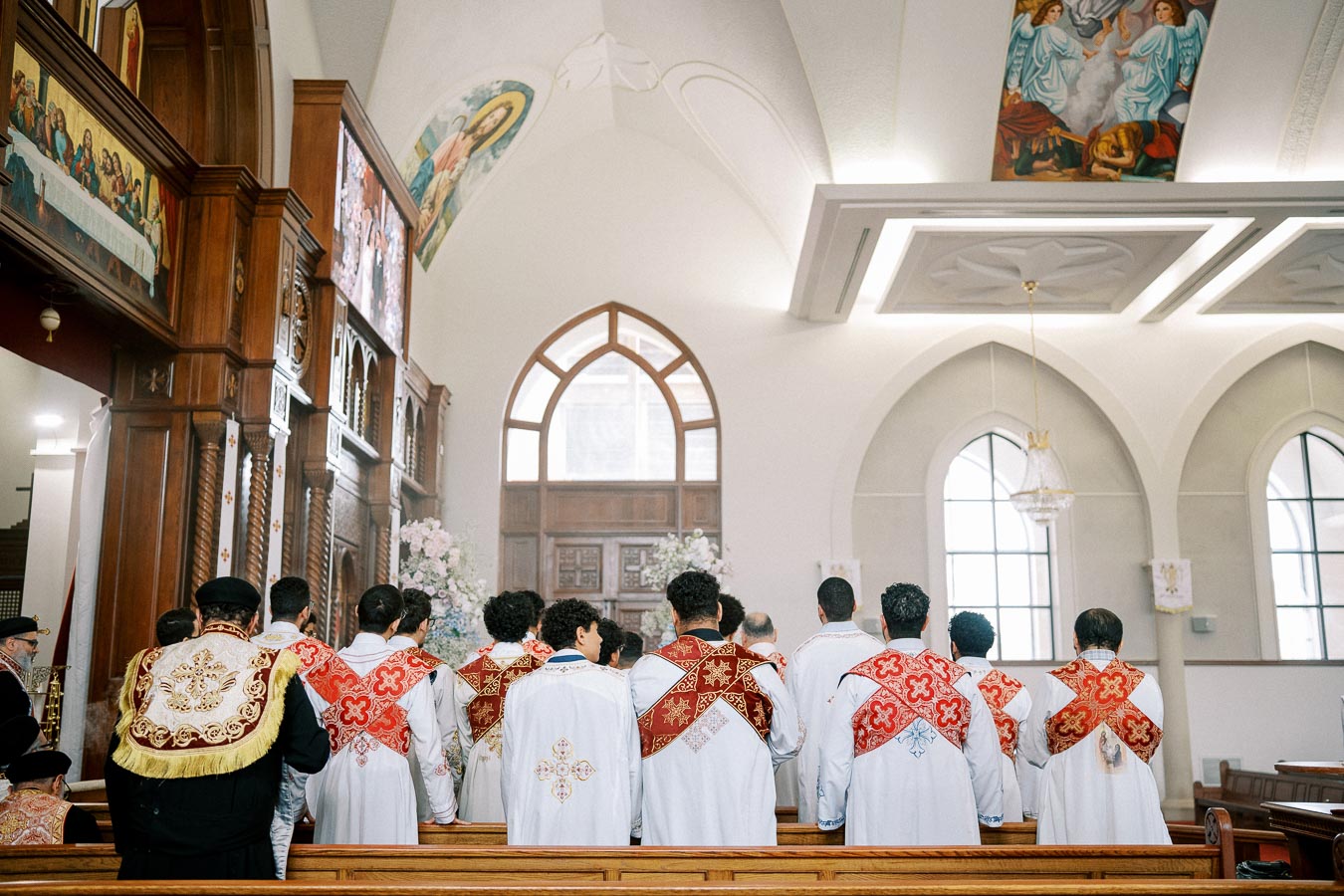 A group of clergy wearing ornate vestments with red and gold designs, standing in prayer inside a church adorned with religious paintings and wooden carvings.