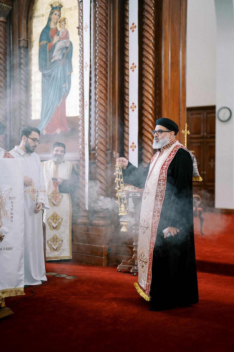 A priest holding an ornate censer during a religious ceremony inside a church, with smoke surrounding him. Marble columns and a painting of Mary holding Jesus are visible in the background. The priest is wearing a black robe with a red and gold stole.