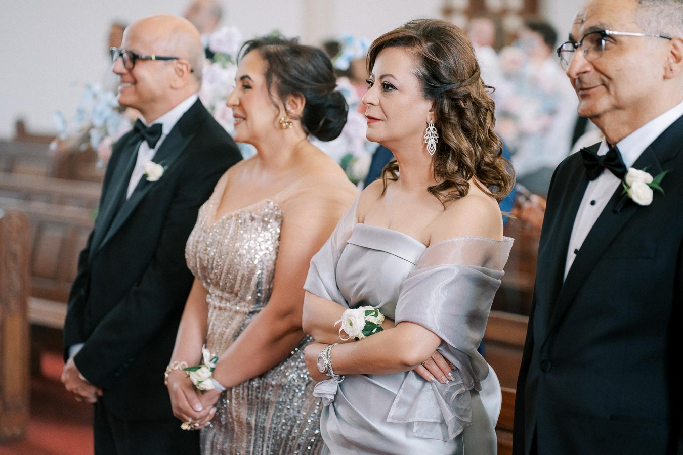 A group of elegantly dressed people standing in a church, with women in formal gowns and men in tuxedos, attentively watching a ceremony.