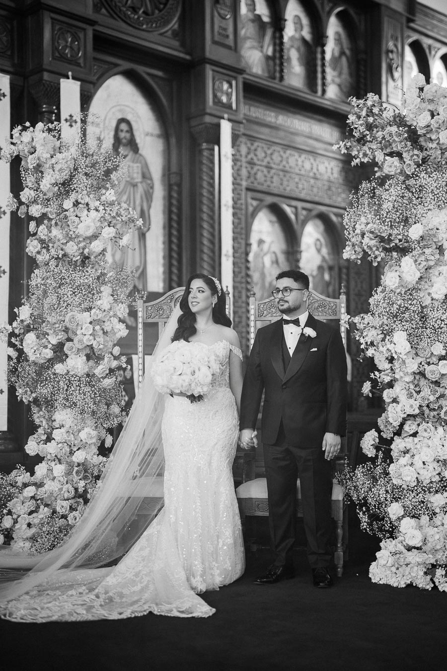 Black and white image of a bride and groom standing at the altar in an ornate church setting, surrounded by elegant floral arrangements. The bride is wearing a lace wedding gown with a long veil, holding a bouquet of flowers, while the groom is in a classic tuxedo.