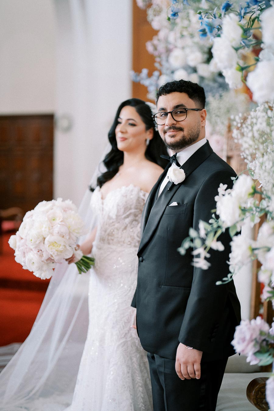 A bride and groom stand together in a beautifully decorated venue. The bride holds a bouquet of white flowers, while the groom wears a black tuxedo with a white boutonnière. Elegant floral arrangements create a romantic setting.