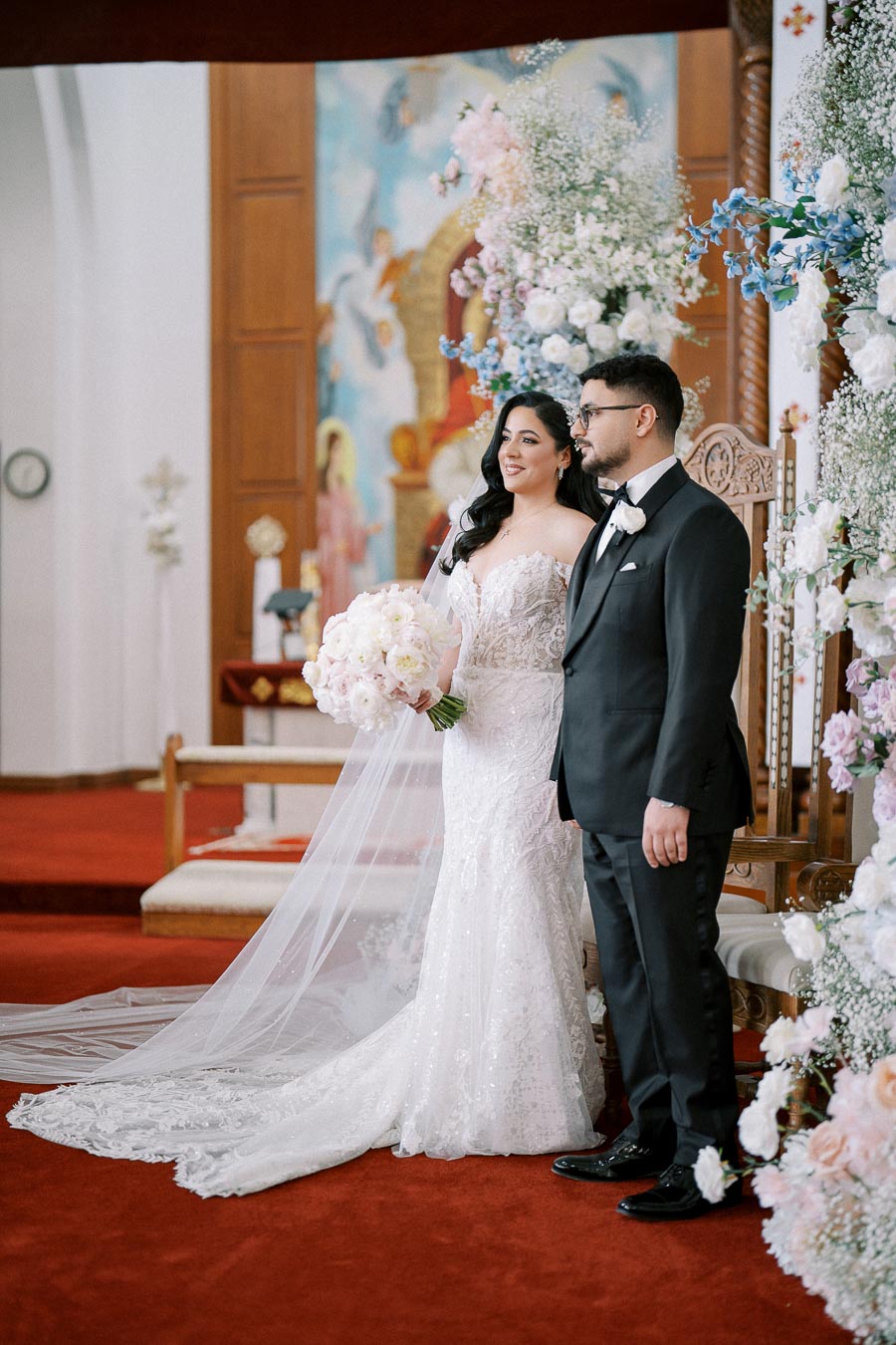 Alt text: Bride and groom standing together at a wedding ceremony, bride holding a bouquet of white flowers, both dressed in formal attire with floral decorations in the background.
