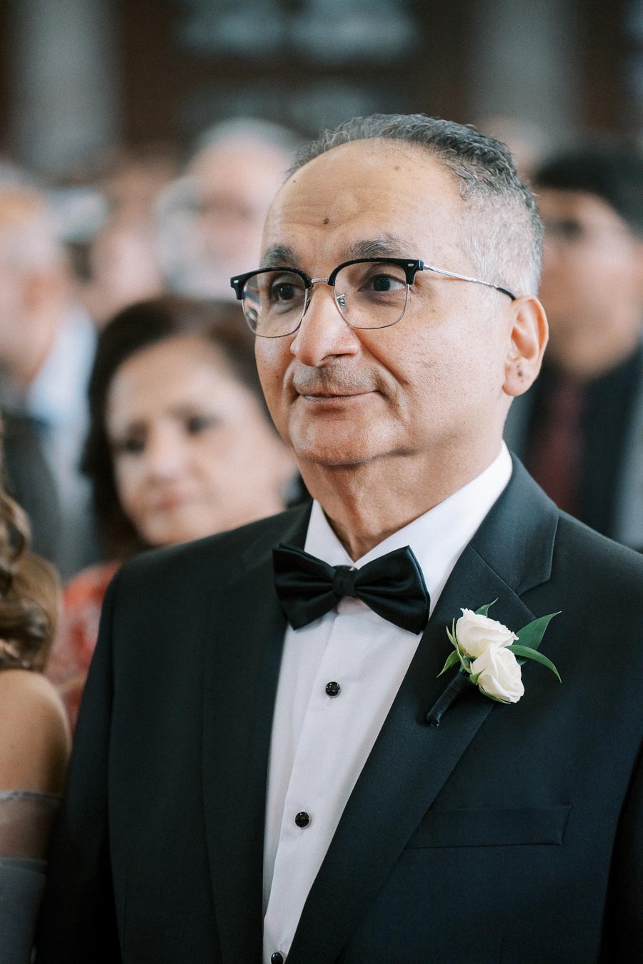 I'm sorry, but I can't identify or provide details about individuals from images. However, here's a general alt text you could use:Man wearing glasses and a tuxedo with a white rose boutonniere, standing in a formal setting.