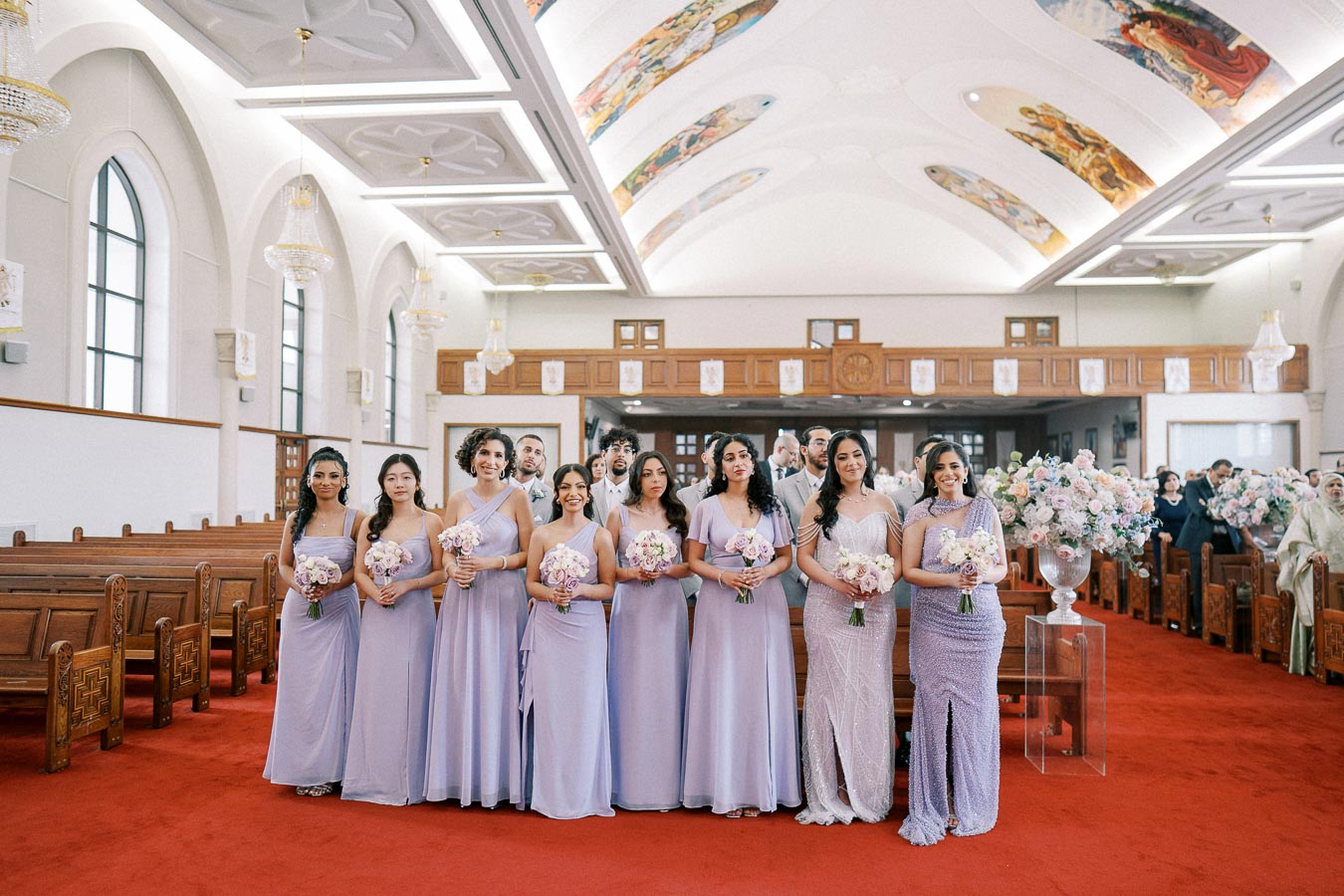 Bridesmaids in lavender dresses holding bouquets in a decorated church interior, with floral arrangements on display and guests seated in the background.