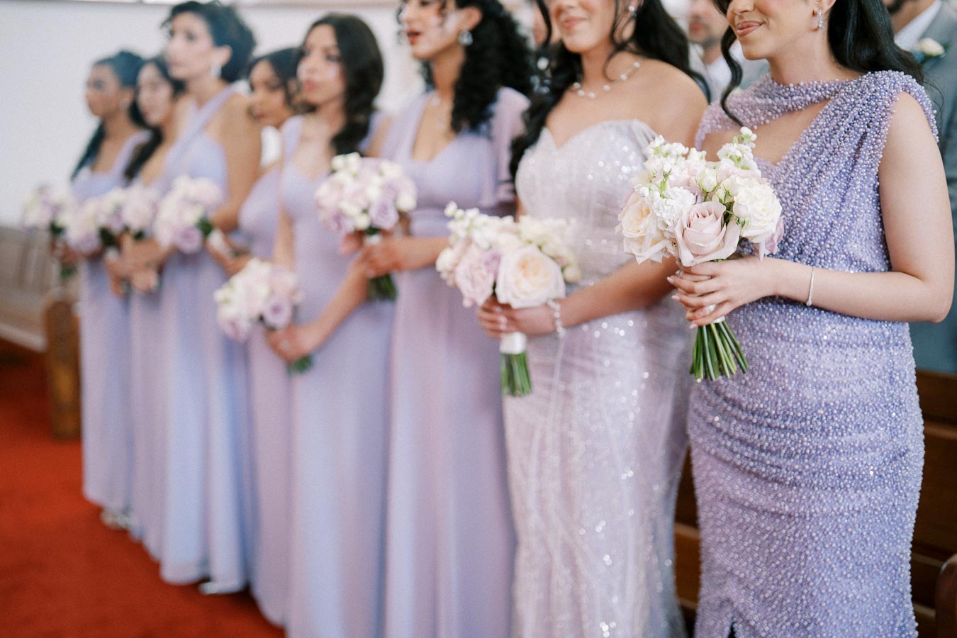 Alt text: Group of bridesmaids in elegant lavender dresses holding pastel flower bouquets standing on a red carpet at a wedding ceremony.