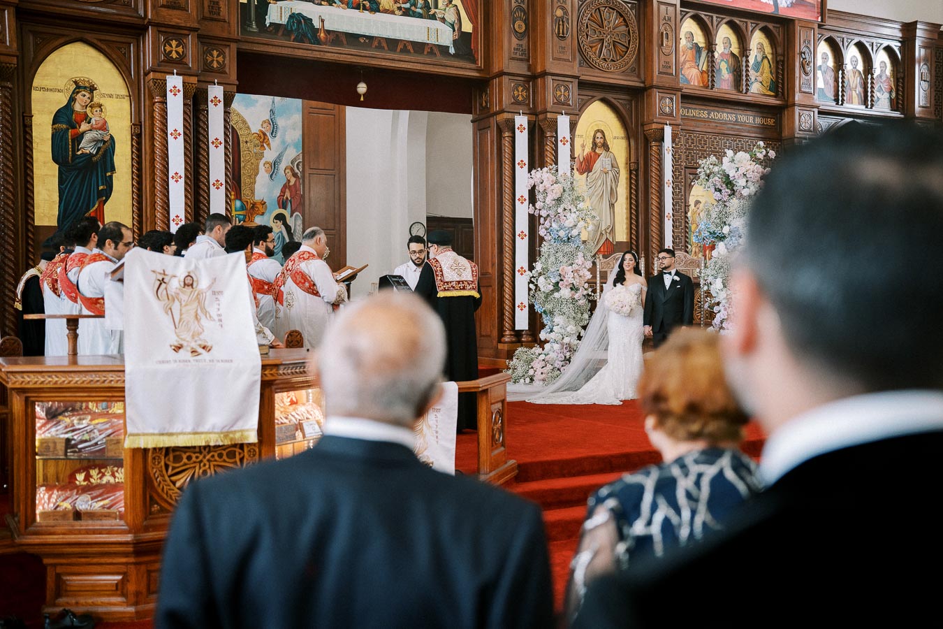 A couple stands at the altar during a traditional church wedding ceremony, surrounded by priests wearing decorative robes, with religious art and floral arrangements adorning the ornate wooden interior.