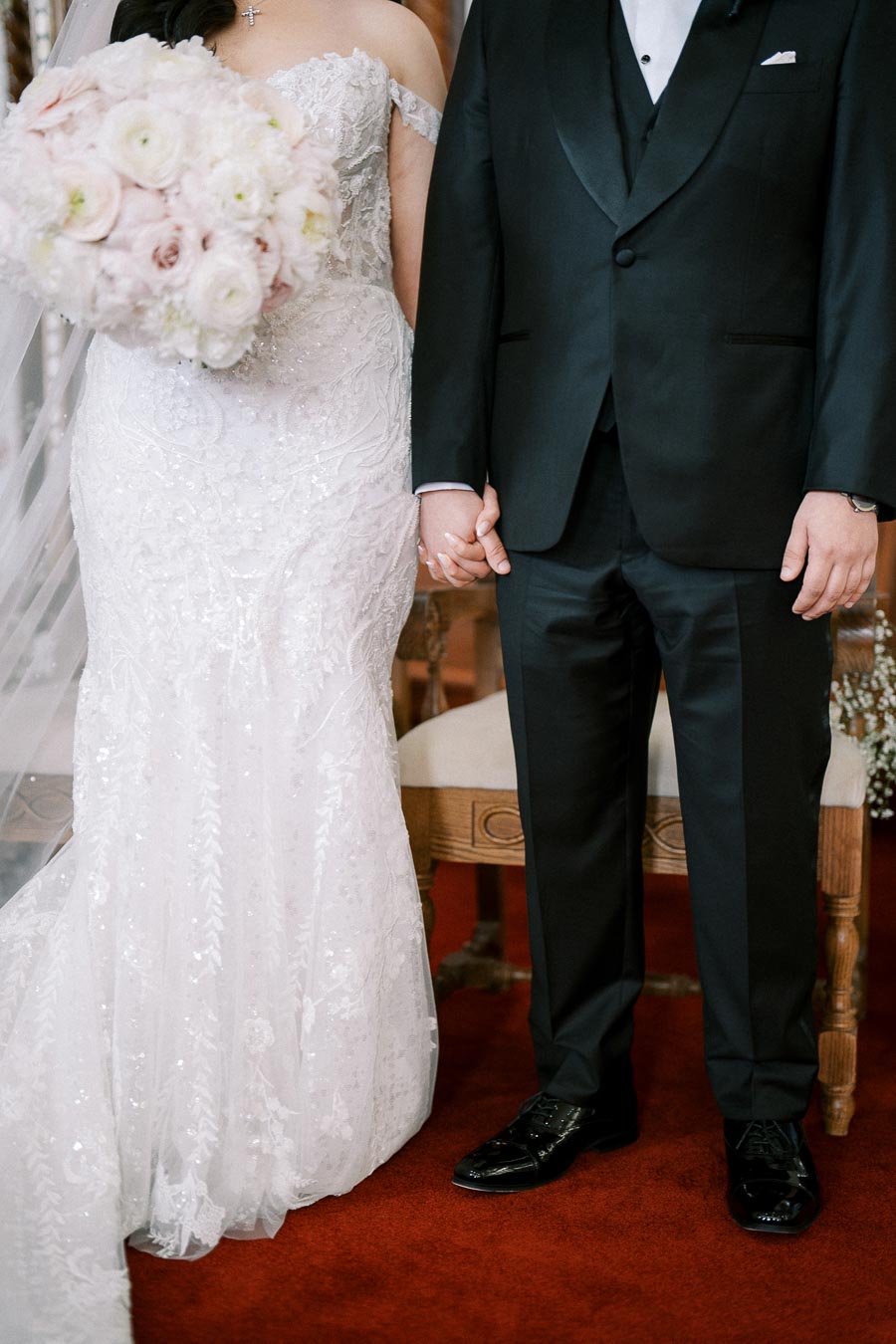 Wedding couple holding hands in elegant attire, featuring a bride in a white lace gown with a bouquet of pale flowers and a groom in a black tuxedo, standing on a red carpet.