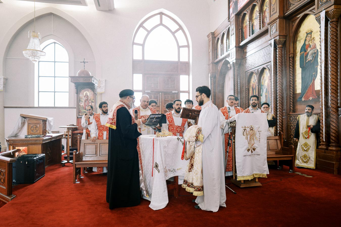 Clergy conducting a religious ceremony in an ornately decorated church with red vestments, iconography, and altar, featuring priests and acolytes participating in a traditional Christian service.