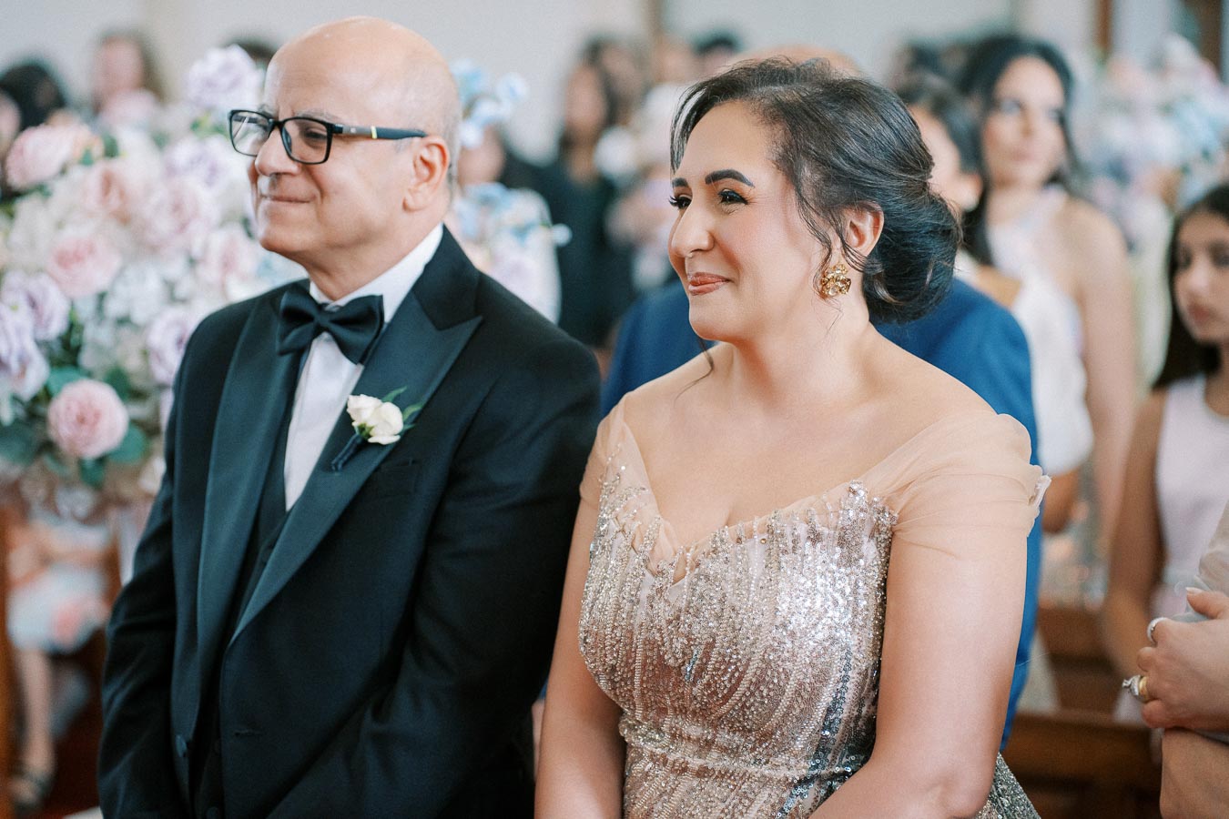 Elderly couple dressed elegantly, sitting together at a wedding ceremony, surrounded by guests and floral decorations.