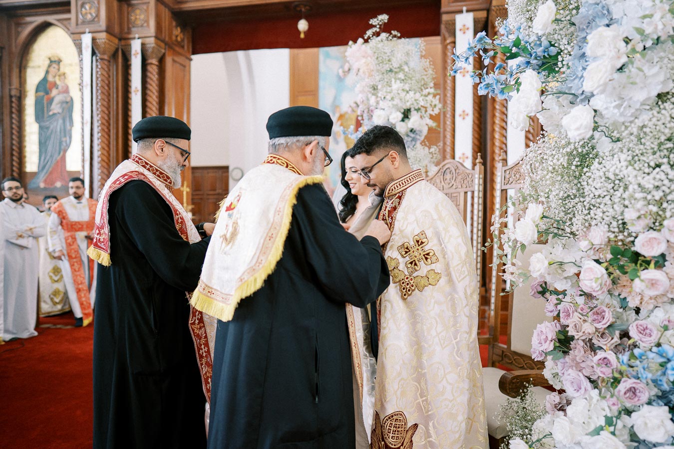 A traditional religious ceremony taking place in a richly decorated church, featuring priests in ornate robes and a couple participating in the ritual, with elaborate floral arrangements and ornate wooden architecture visible in the background.