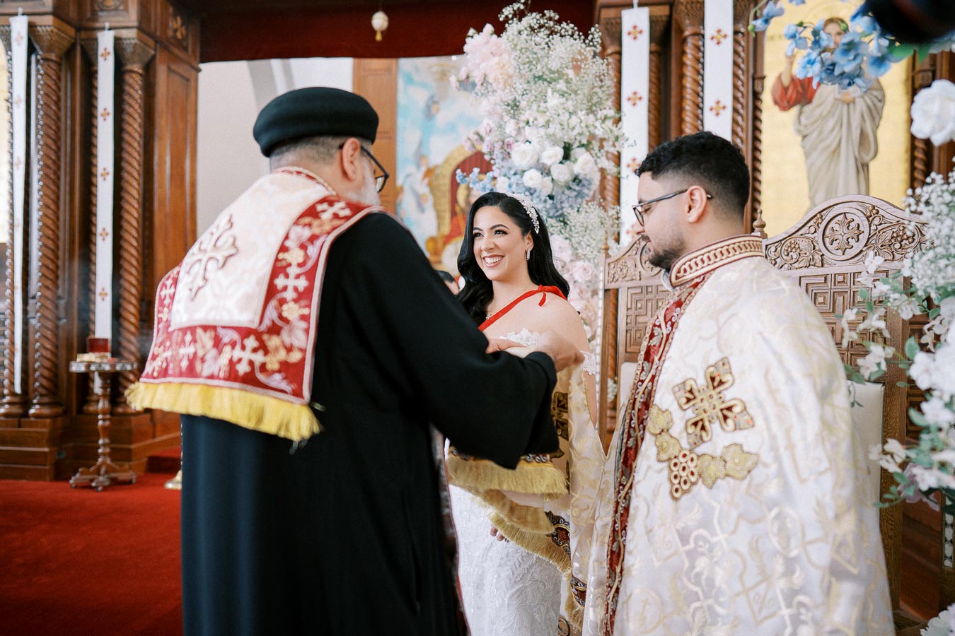 A joyful bride and groom stand before a priest during a traditional wedding ceremony, adorned in ornate attire, set in a beautifully decorated church with floral arrangements and religious icons.