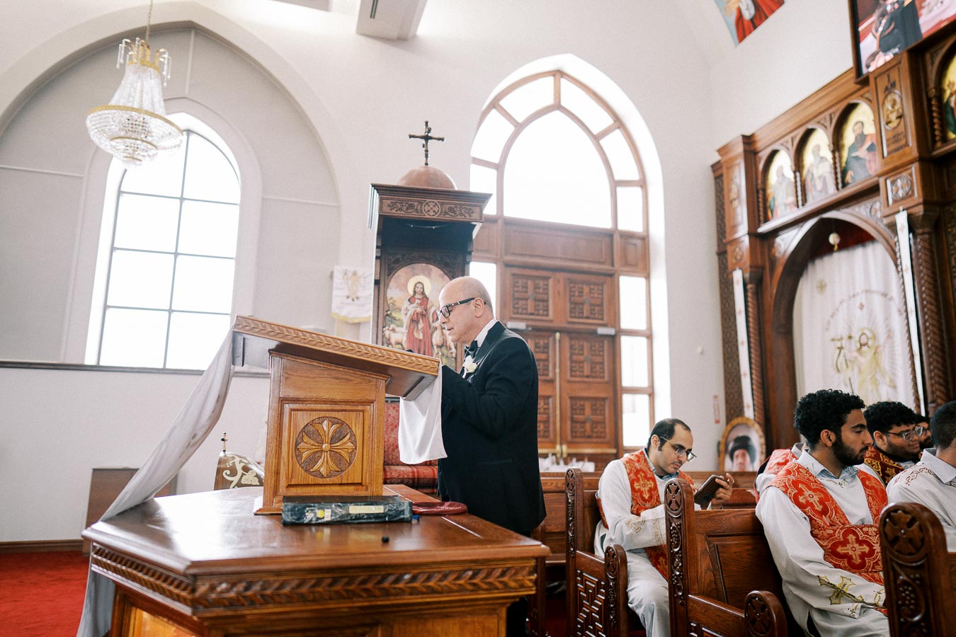 A man stands at a wooden lectern inside a beautifully decorated church, reading from a book. The church features large arched windows, ornate woodwork, and religious imagery. In the background, religious figures in white and red robes are seated, and a large chandelier hangs from the ceiling.