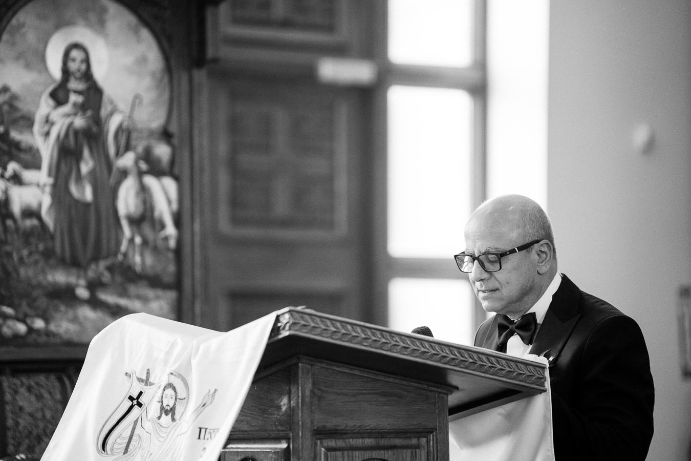 A man in a suit and glasses stands at a pulpit in a church, speaking or presenting. In the background, a religious painting featuring a shepherd and sheep is visible. The scene is in black and white, conveying a formal and solemn atmosphere.