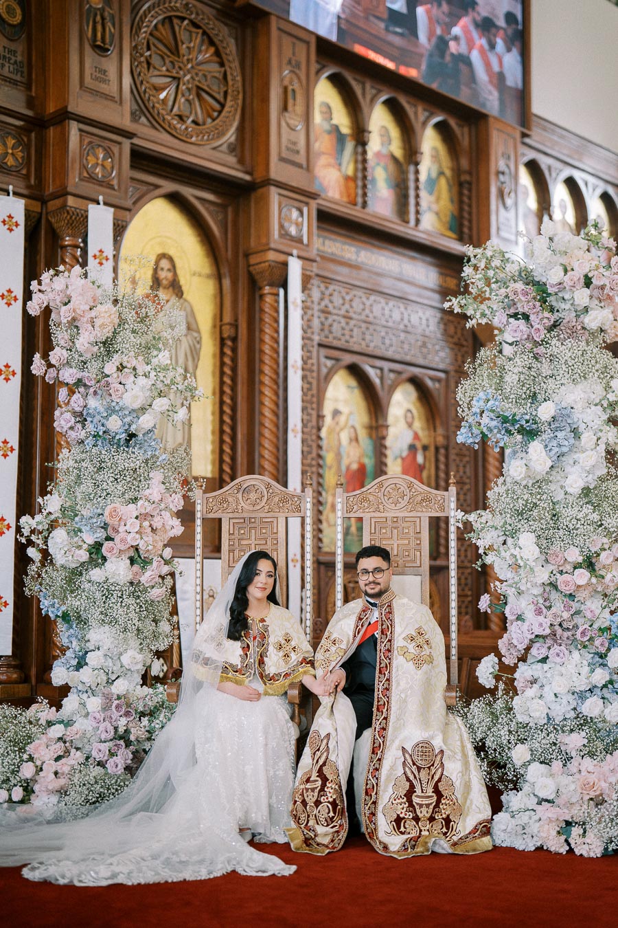 A bride and groom in ornate traditional attire sit on elaborately carved chairs, surrounded by lush floral arrangements in a beautifully decorated church setting.