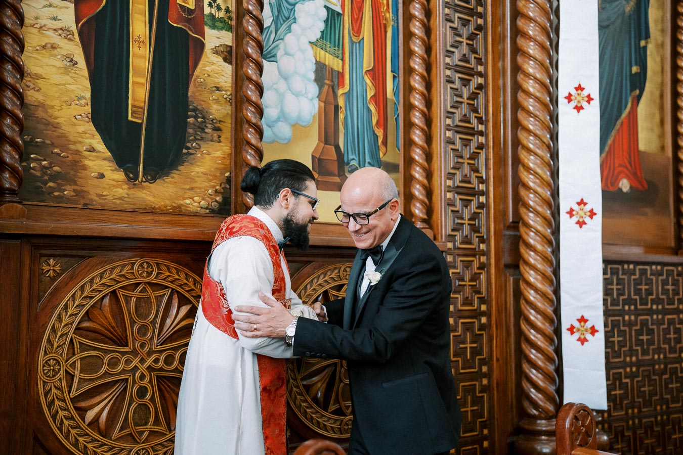 A priest and a man in a suit share a warm greeting inside a richly decorated church, adorned with intricate wooden carvings and religious artwork.