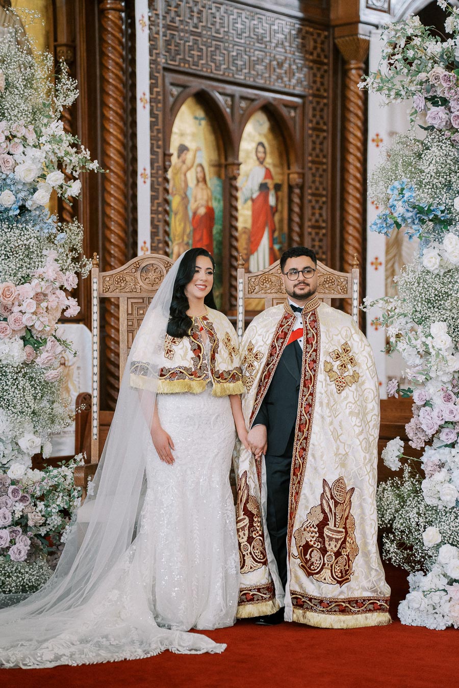 Bride and groom in traditional ornate attire at a church, surrounded by floral arrangements, standing on a red carpet.