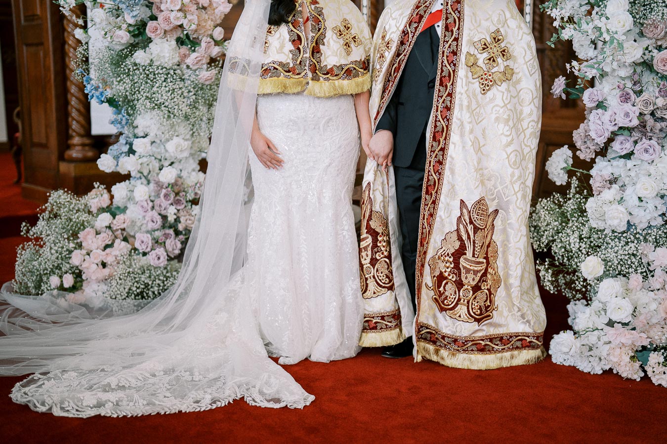 Elegant wedding photo featuring a bride and groom in traditional embroidered garments, surrounded by a floral arch with pastel roses and baby's breath, on a red carpet.