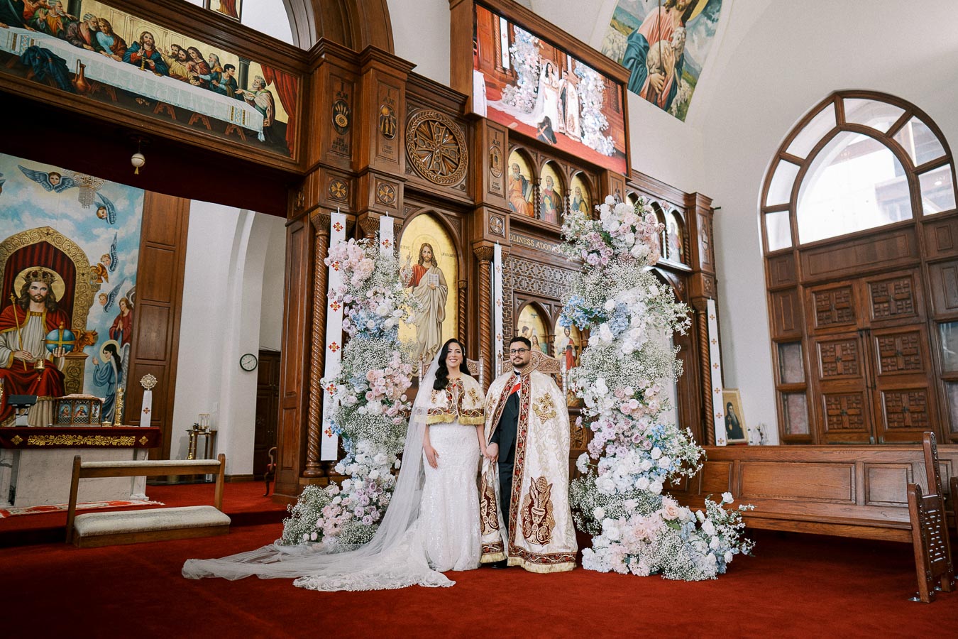 Bride and groom in traditional attire standing in a beautifully decorated church with floral arrangements and religious artwork in the background, celebrating their wedding ceremony.