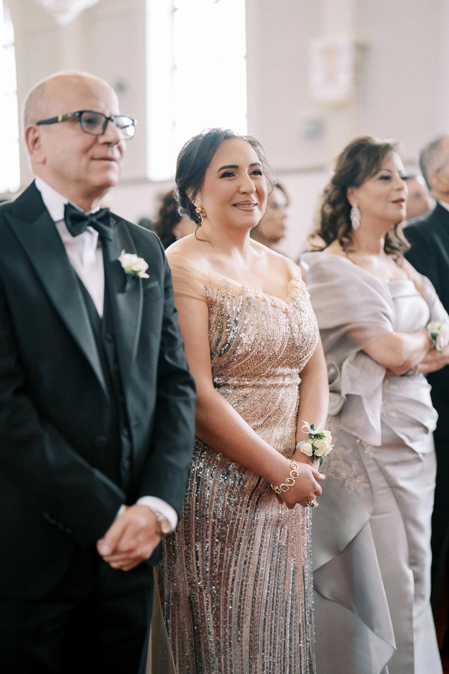Elegant group of mature adults dressed in formal attire attending a special occasion in a well-lit venue, with the man in a black tuxedo and two women in glittering evening gowns.