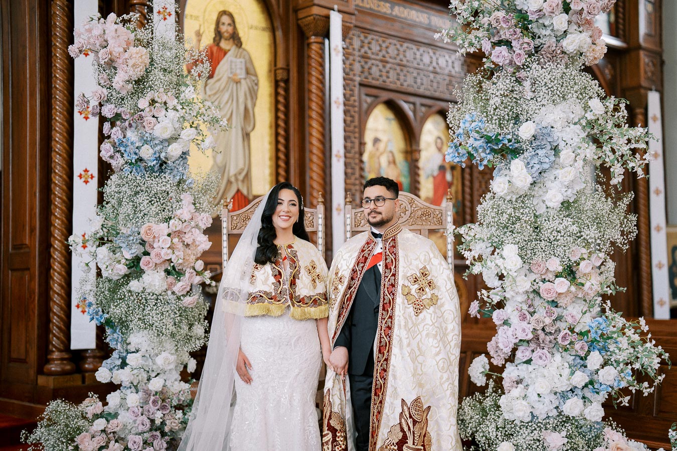 A bride and groom in ornate traditional attire holding hands during their wedding ceremony in a beautifully decorated church with floral arrangements and religious icons.