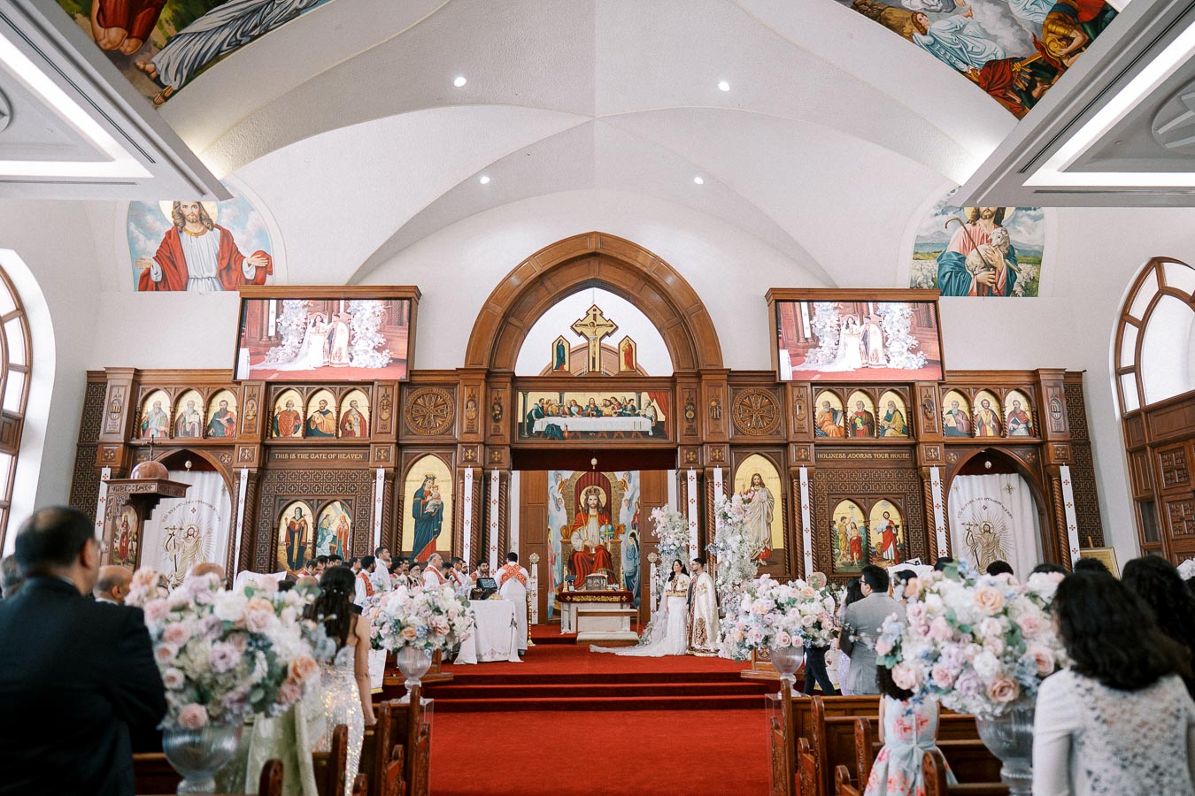 Interior of a beautifully decorated church during a wedding ceremony. The ornate altar is adorned with religious artwork and floral arrangements. Guests are seated on both sides of the aisle, witnessing the ceremony. The ceiling features intricate patterns and religious imagery.