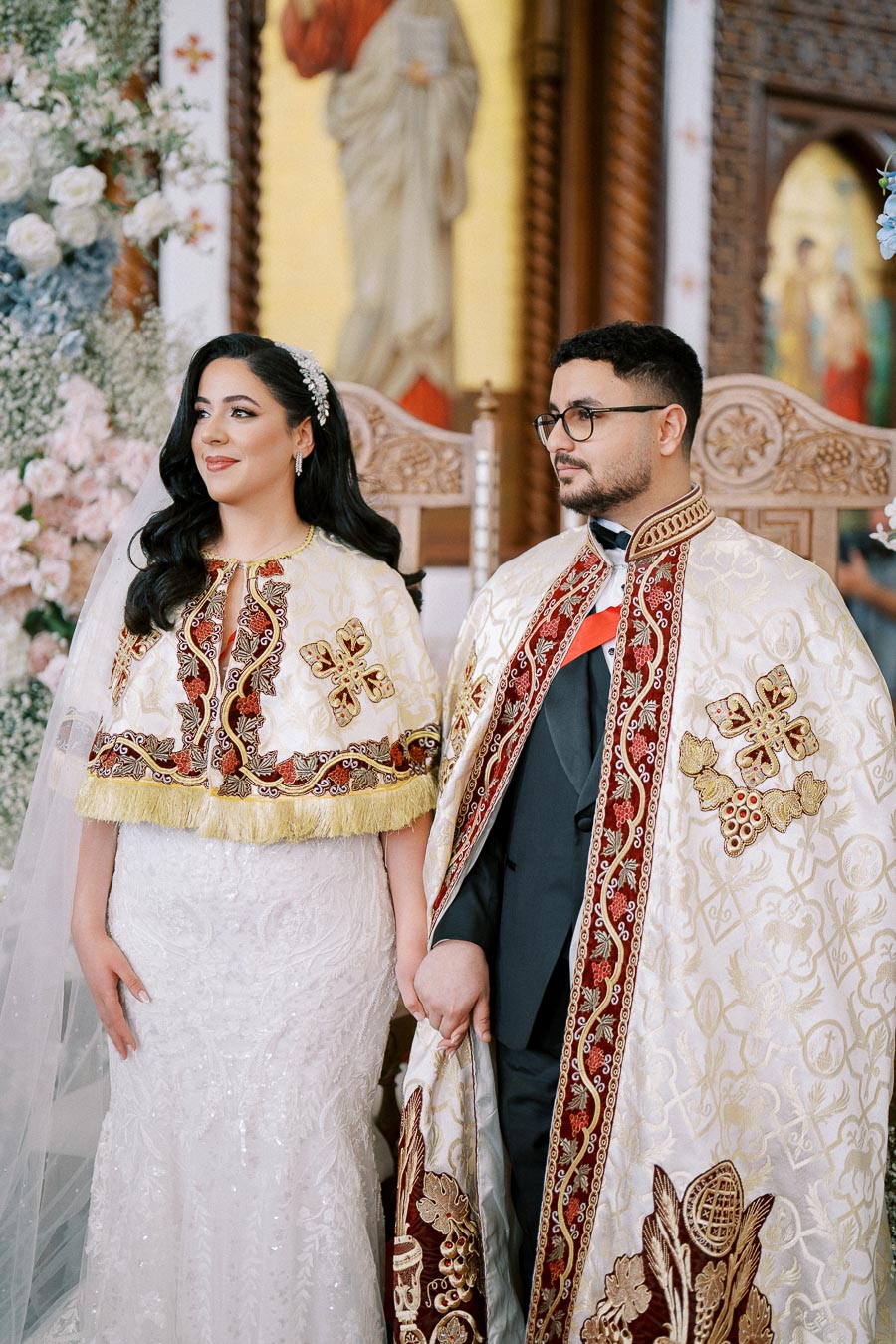 Traditional wedding ceremony with bride and groom wearing ornate gold and red embroidered robes, standing in a beautifully decorated church setting.
