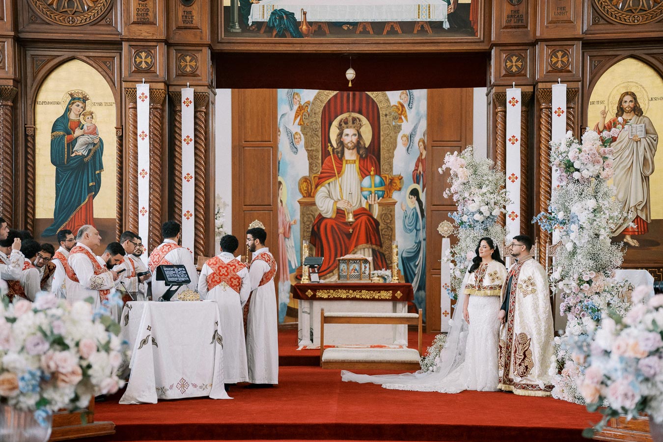 A couple getting married in an ornate church with priests in white and red robes, surrounded by floral decorations and religious iconography.