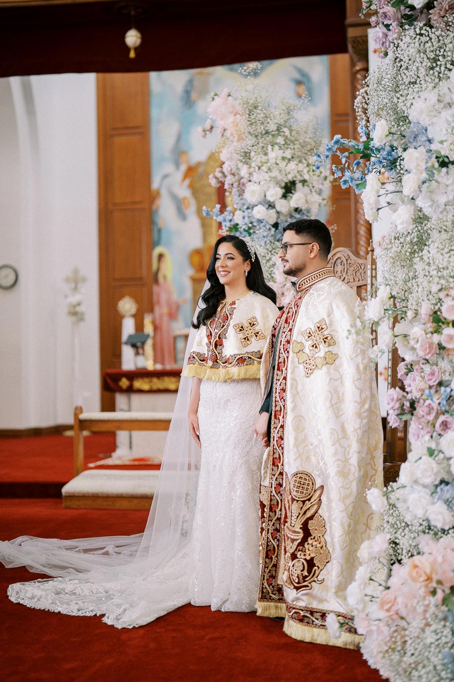 A bride and groom in traditional ornate garments stand together at an altar, surrounded by vibrant floral arrangements, during a wedding ceremony in an ornately decorated church interior.