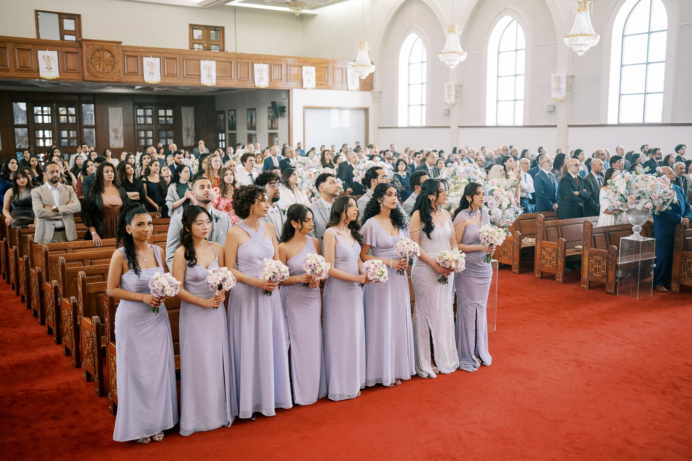 A group of bridesmaids in lavender dresses holding flower bouquets stand in a church with a gathered audience during a wedding ceremony.