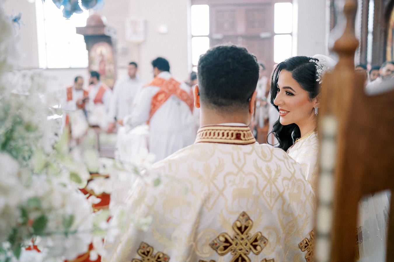 A newlywed couple in ornate traditional wedding attire smiles during a religious ceremony inside a beautifully decorated church, with priests in red sashes in the background.