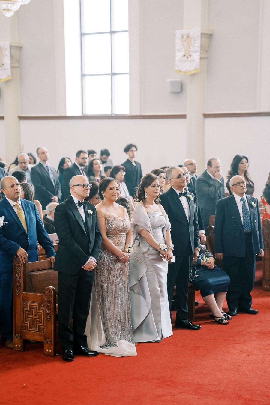 A formal gathering at a wedding ceremony with elegantly dressed guests standing and seated in a church, showcasing a joyful and celebratory atmosphere.