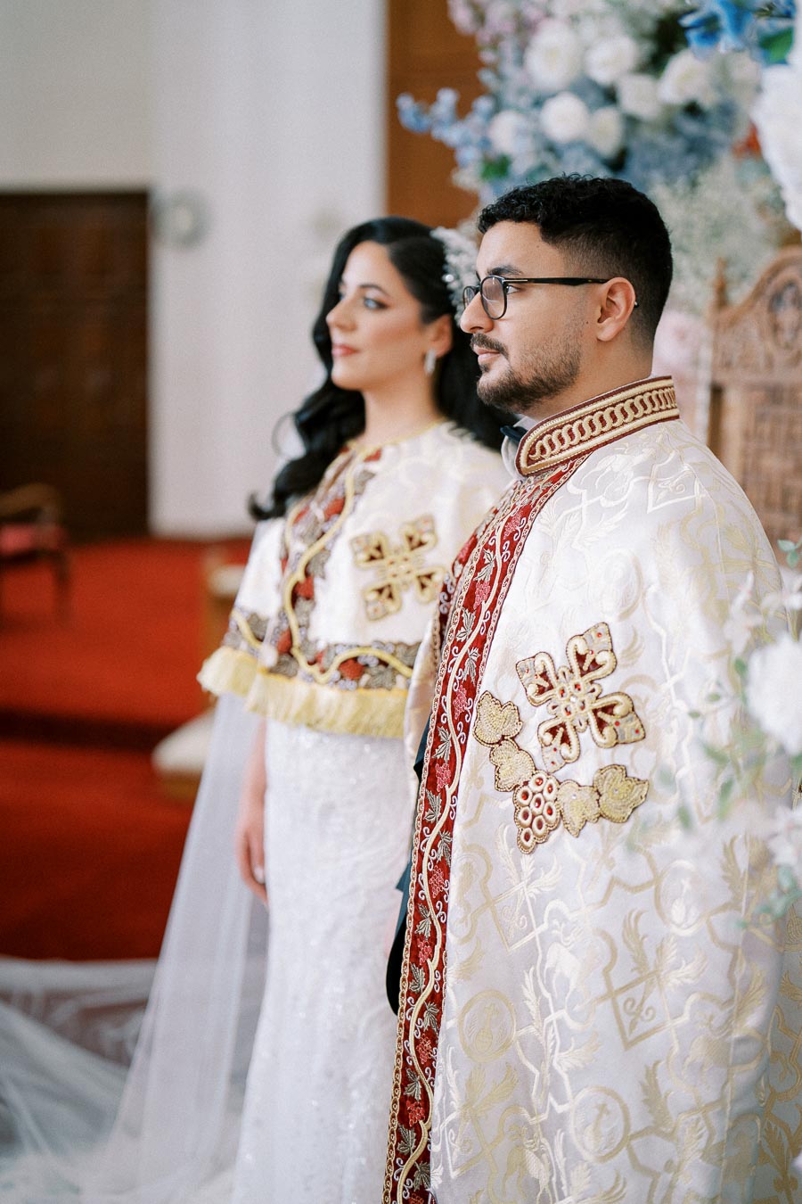 Couple in traditional ceremonial attire stands in a beautifully decorated hall with floral arrangements, capturing a significant cultural event.
