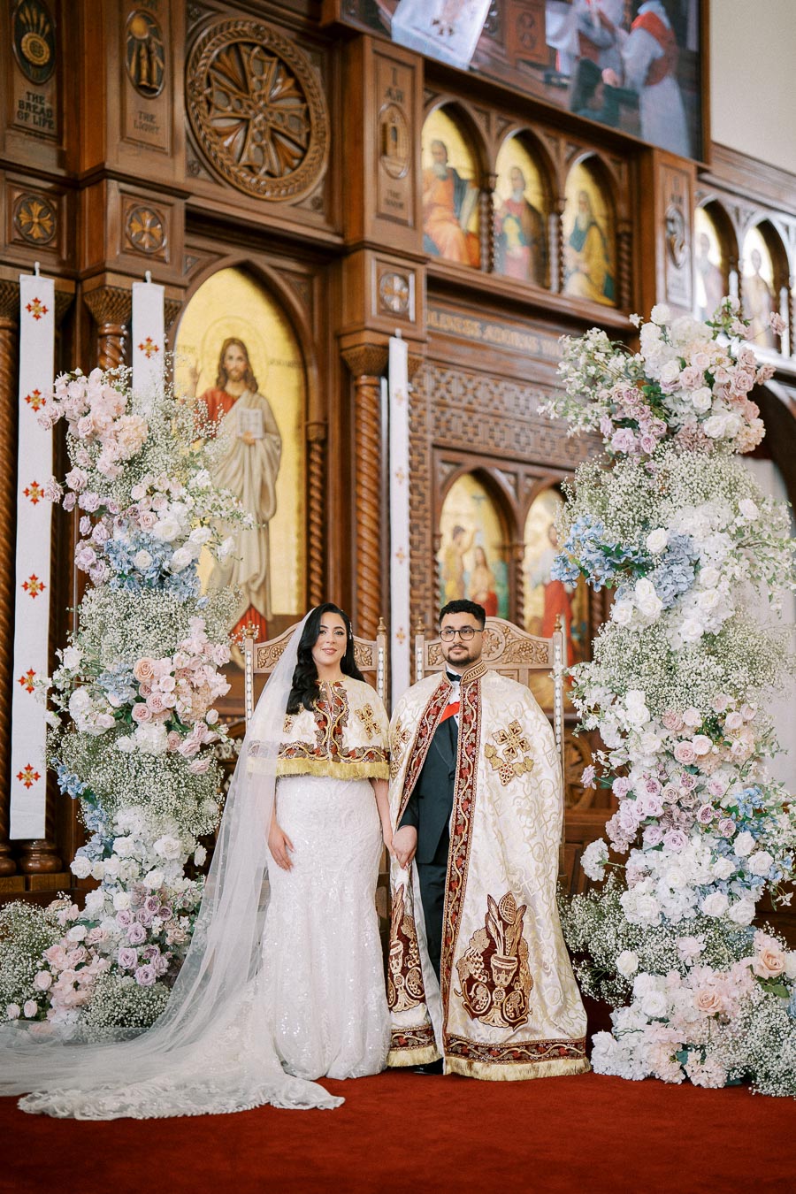 Alt text: Bride and groom in traditional attire standing in a beautifully decorated church with floral arrangements and ornate religious icons. The couple is dressed in ceremonial garments, holding hands, against a backdrop of wooden carvings and colorful religious artwork.